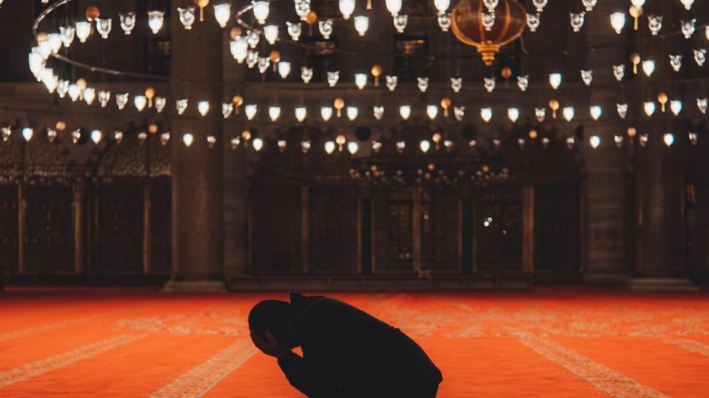 A Muslim man kneeling in silent prayer inside a beautifully illuminated mosque